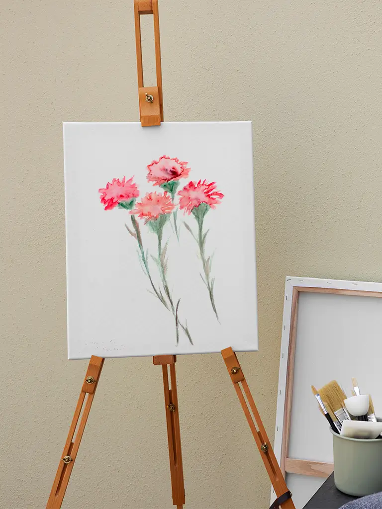Pintura en acuarela de flores rosadas sobre lienzo blanco, colocado en un caballete de madera junto a pinceles y materiales de arte.