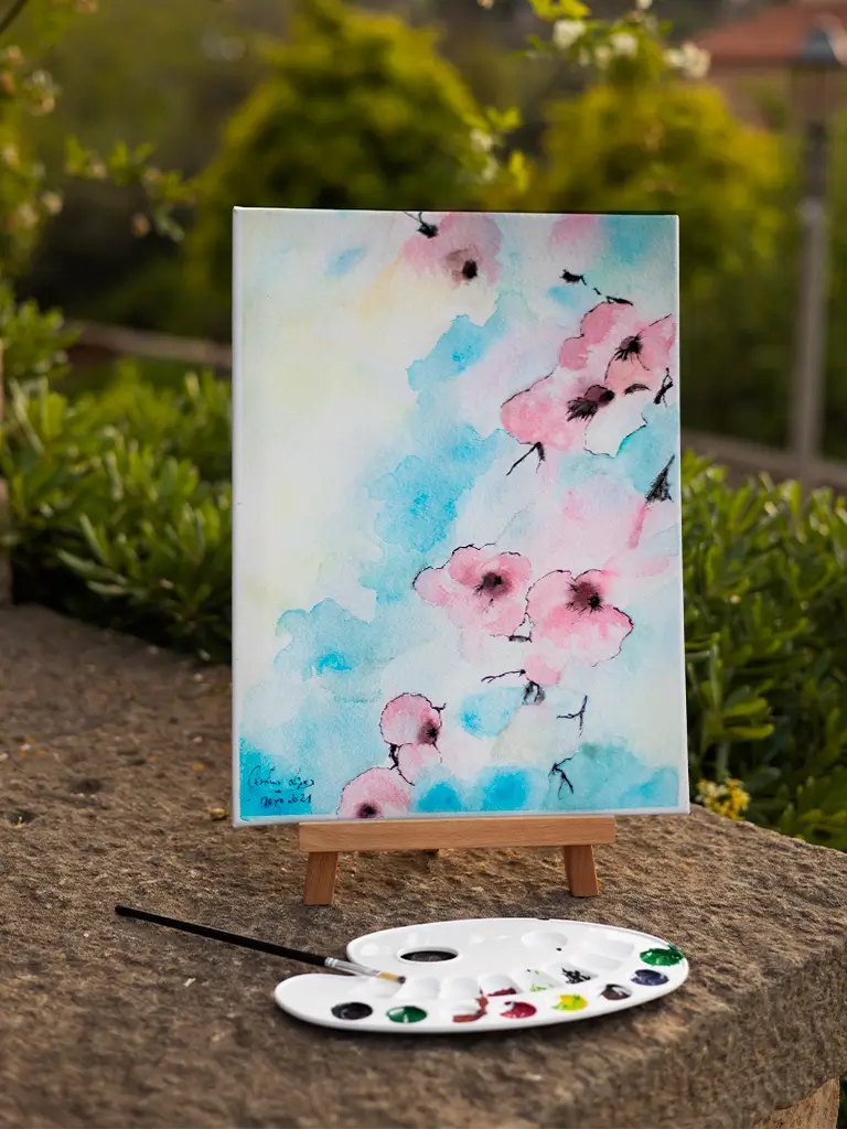Pintura en acuarela de flores rosadas sobre fondo azul, colocada en un caballete de madera al aire libre junto a una paleta de pintura.