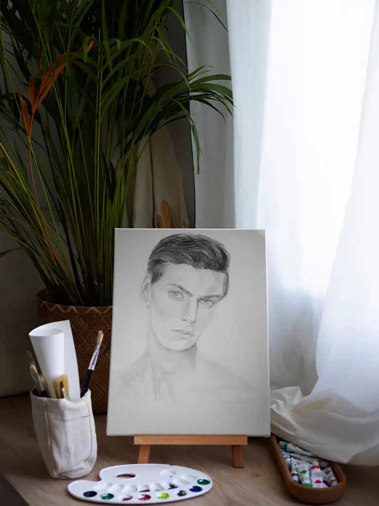 Retrato a lápiz de un hombre joven sobre caballete, rodeado de materiales de pintura y una planta, junto a una ventana con cortinas blancas.