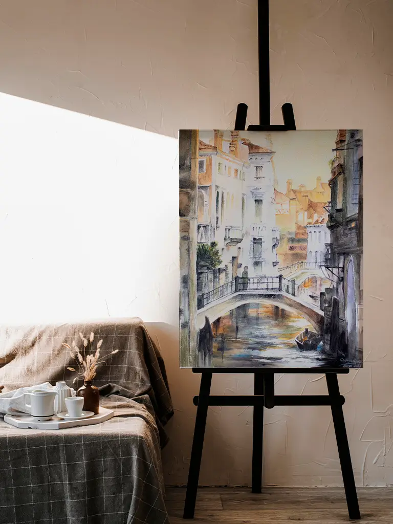 Cuadro en acuarela de una ciudad con canal y puente sobre caballete negro, junto a sofá con bandeja de té y decoración acogedora.