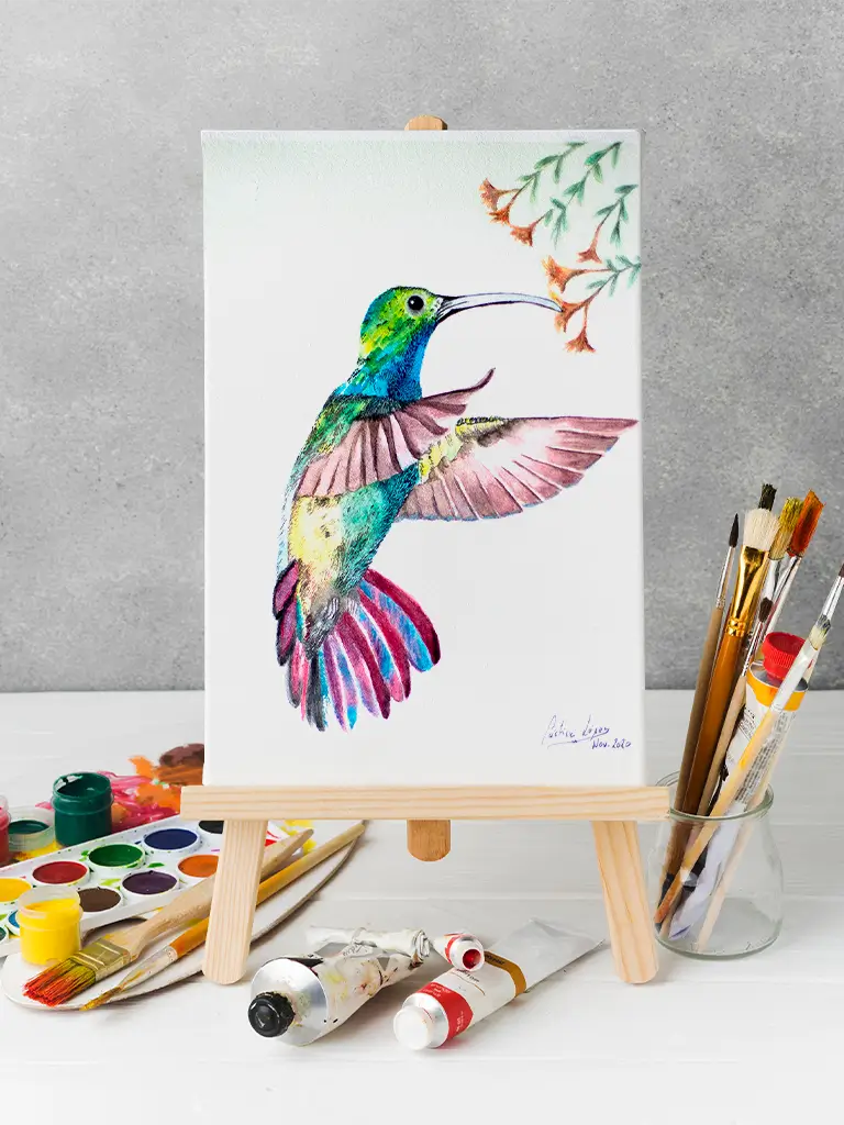 Pintura en acuarela de un colibrí colorido alimentándose de una flor, sobre un lienzo en un caballete, rodeado de pinceles y tubos de pintura.